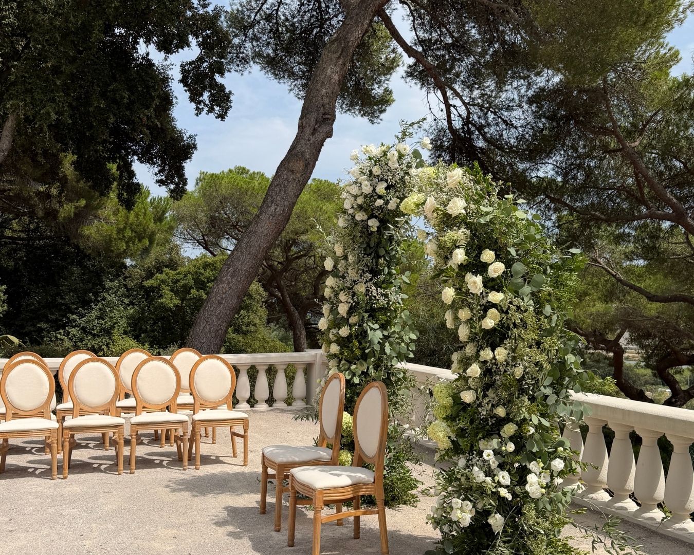 Allée de cérémonie avec chaises en bois et arche de fleurs blanches dans un jardin méditerranéen